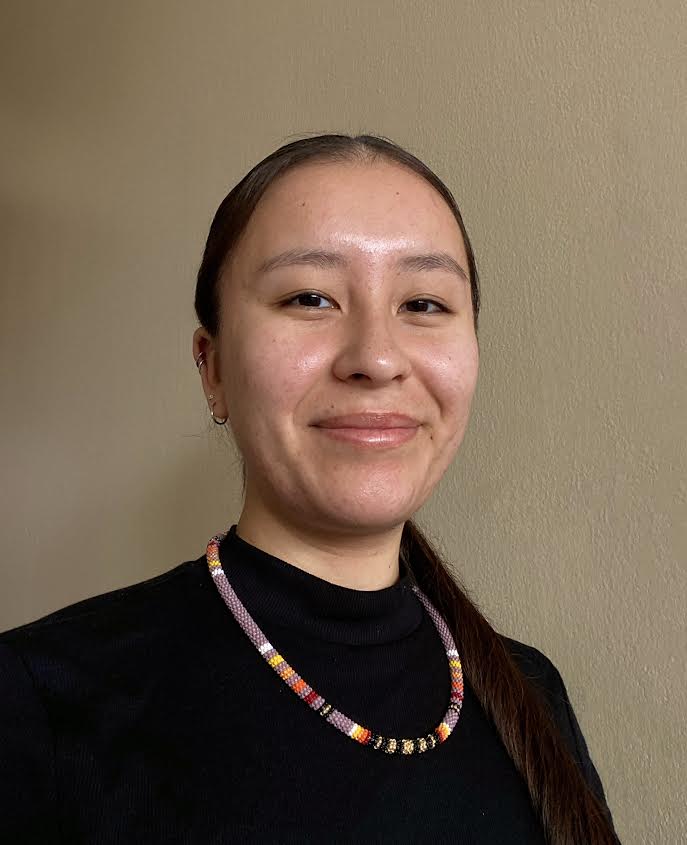 Portrait of Joleece Pecore. Joleece smiles against a neutral-colored wall. She is wearing a black top and a beaded Native necklace. 
