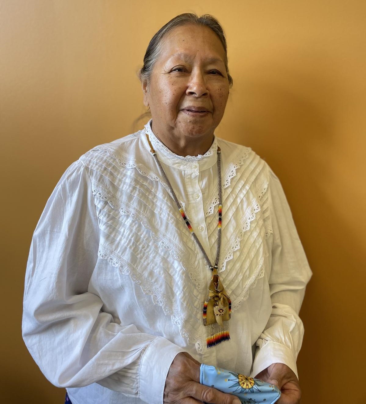headshot of Elder in Residence (2023) Nelda Goodman. Nelda is pictured against an orange wall wearing a white cloth shirt.She is smiling and holds a patterned cloth. She is wearing a traditional medicine bag around her neck.