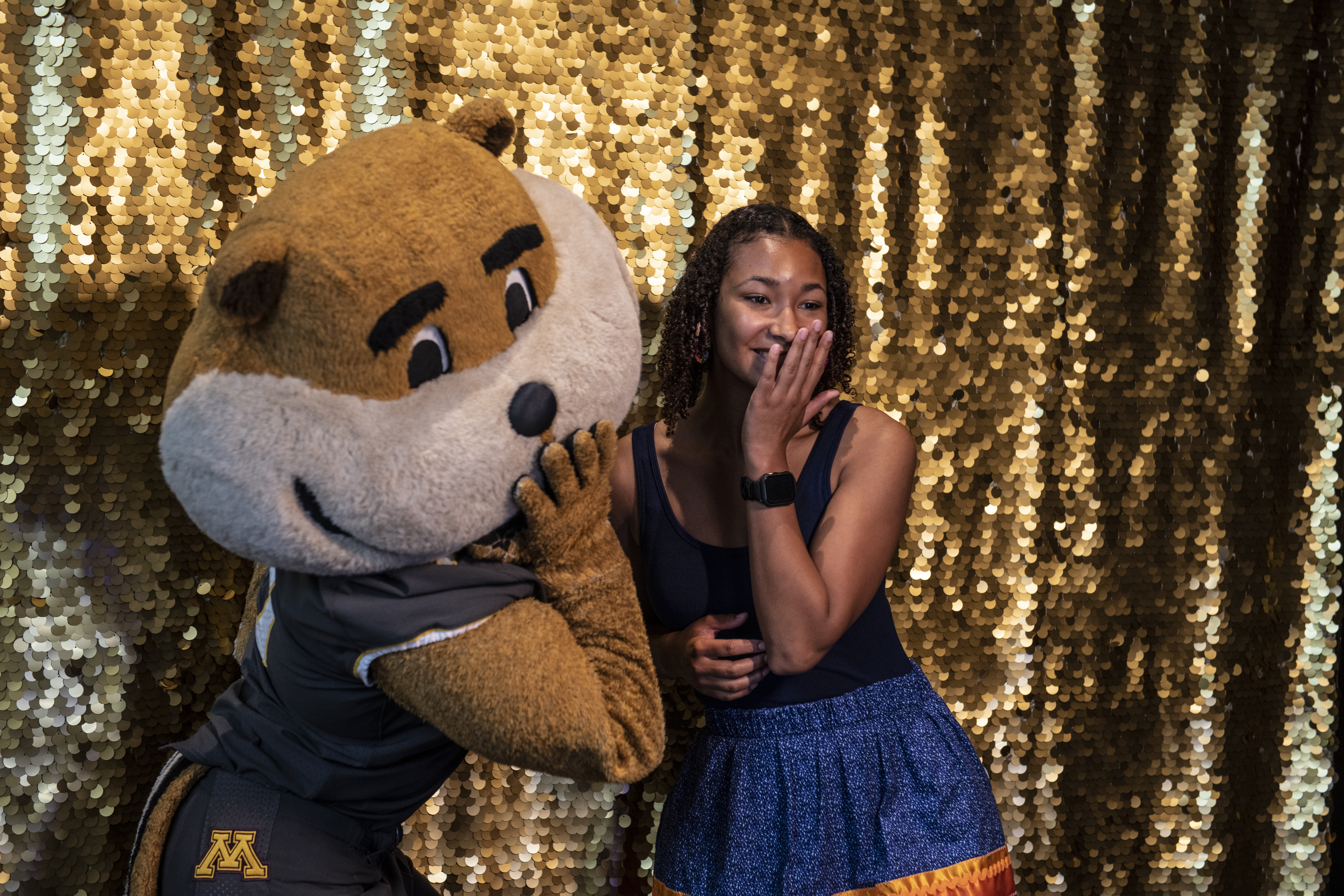 A Native student poses for a picture with Goldy Gopher at the annual N&I KO Program. The student and Goldy pose with their hands over their mouths. The student is wearing a blue ribbon skirt, and Goldy is wearing the UMN football uniform