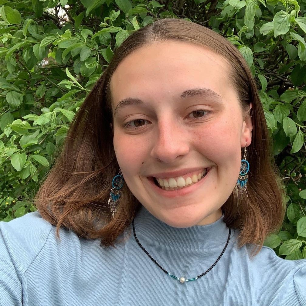 Taylor Fairbank's staff headshot. Taylor is wearing Native jewelry, and smiles in front of a green foliage background. She is wearing earrings, and a necklace. She has shoulder-length brown hair.