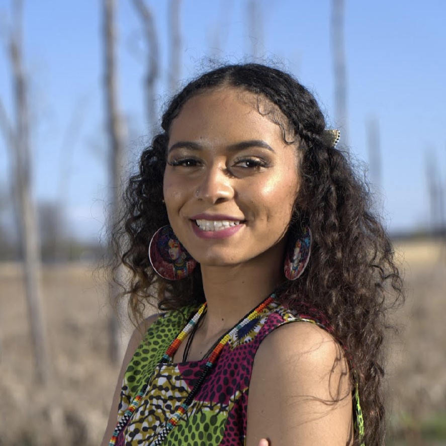 Sierra Charwood's official headshot: Sierra stands in front of a blurred grassy wooded background. She has curly long hair. She is wearing a beaded necklace, and decorative earrings with a woman painted on them. She is smiling. 