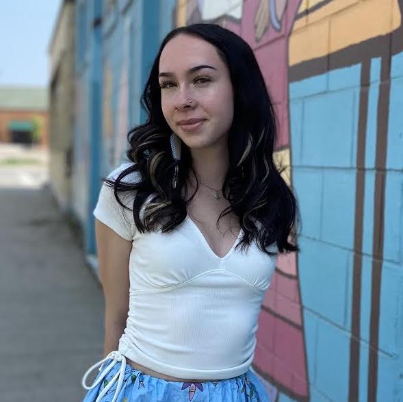 Ava Hartwell's official headshot. She has dark shoulder-length hair, and wears a white top with a blue Native ribbon skirt. She holds her hands behind her back, and smiles in front of a blue mural painted on a wall in the background