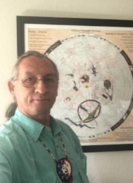Jim Rock's official headshot. He stands in front of his D(L)akota Star Constellation Guide (map/graphic). He is wearing a teal shirt, and a beaded Native medallion of a sun and moon around his neck. He has glasses, and wears his hair in a ponytail.